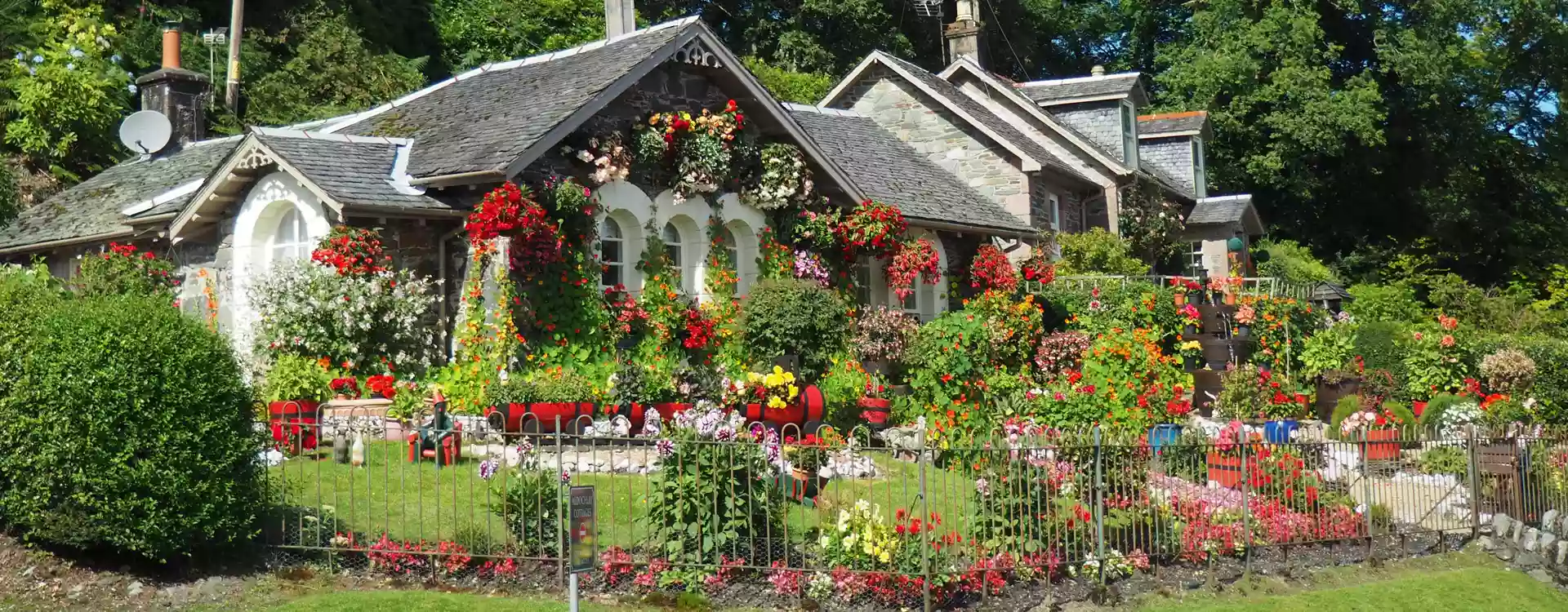 Natursteinhaus mit Beeten, die mit bunten Blumen bepflanz sind und einem satt grünen Rasen mit Edelstahlzaun
