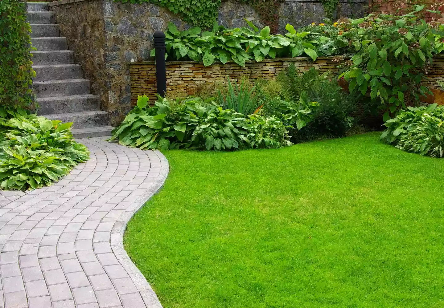 Gepflasterter Steinweg aus weißen Steinen führt zu einem Hausaufgang, vorbei an einer Wiese aus Rollrasen mit einer romantischen Natursteinmauer und Sträuchern