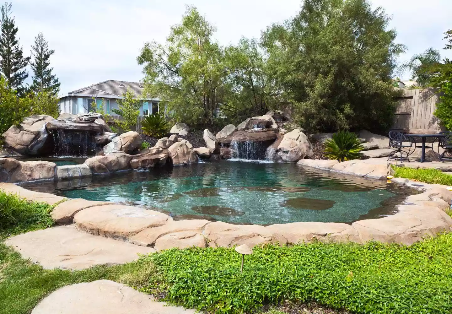 Garten- und Landschaftsbau Schwimmteich mit Natursteinelementen und Wasserfall in einem Garten mit Wiese, Wildpflanzen und Baumbestand